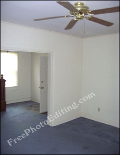 Photograph of an empty room with bare white walls and somewhat damaged carpetting. The room has a decorative ceiling fan.